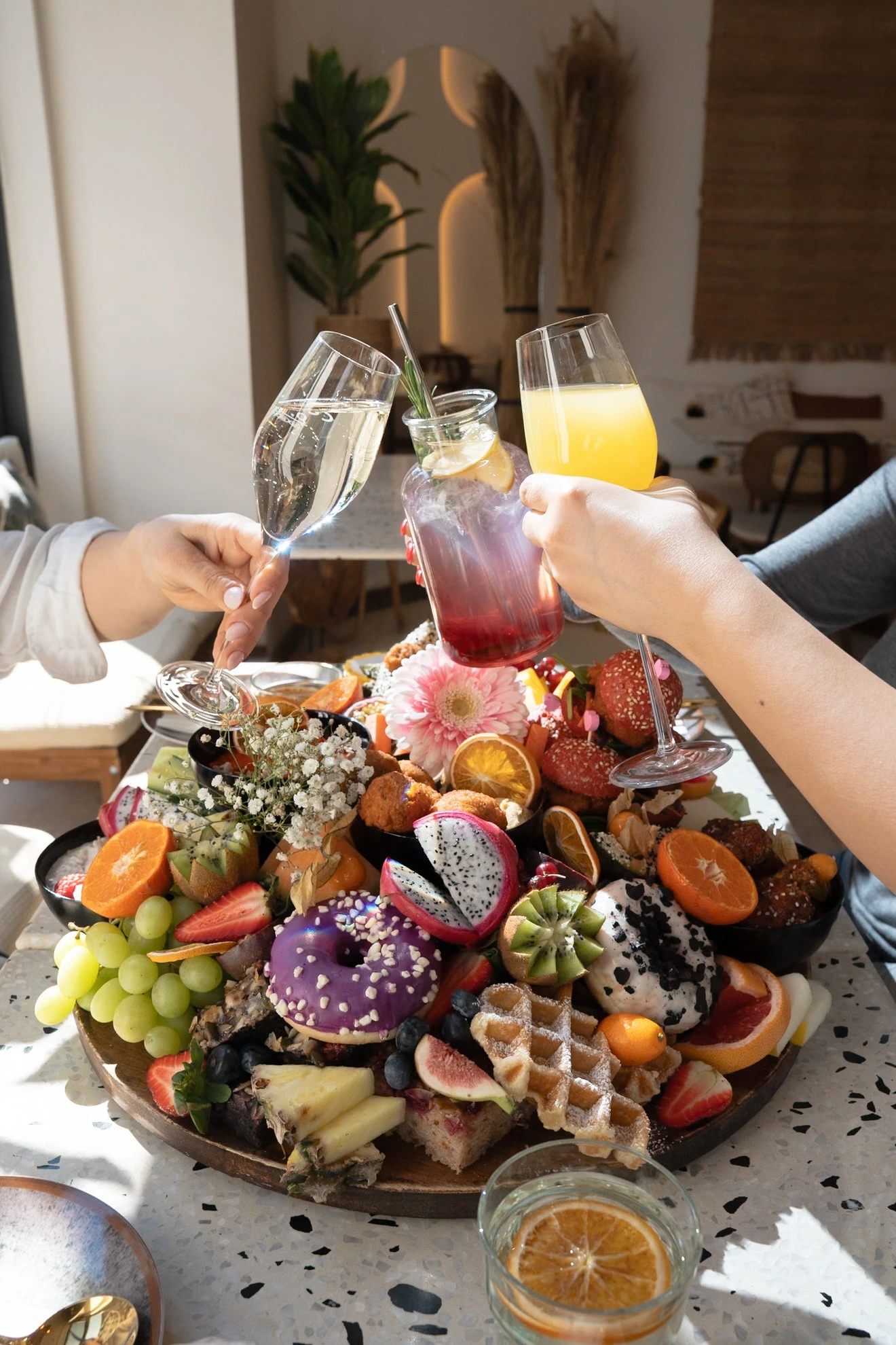 3 Hände, die mit verschiedenen Getränken über einer vegetarischen Brunchplatte anstoßen